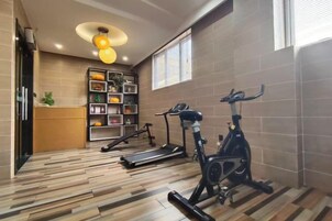 Un gimnasio con equipo de ejercicio, piso de madera, estante en la pared con objetos y una ventana.