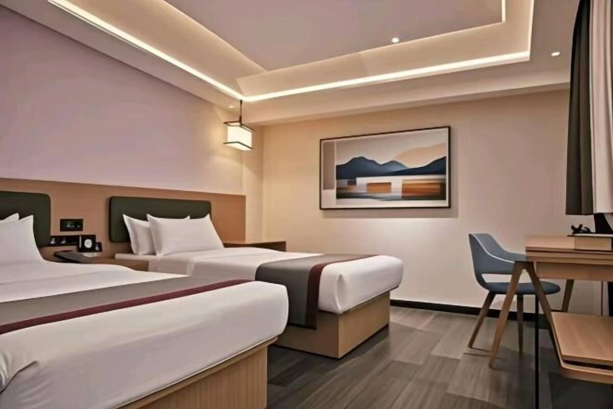 A hotel room with two beds, a desk, a chair, a painting on the wall, and a clock on the nightstand.