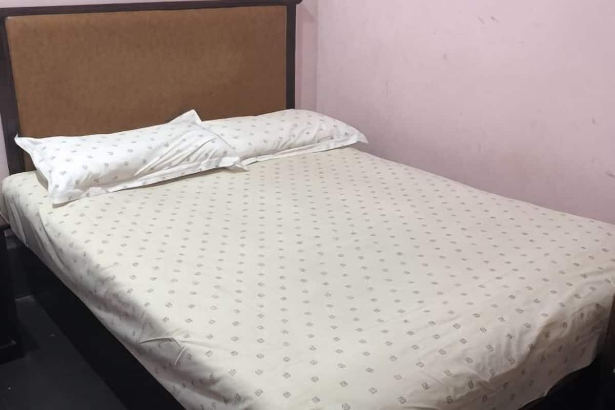 A single bed with a patterned bedspread and pillows, a wooden headboard, and a plain wall.