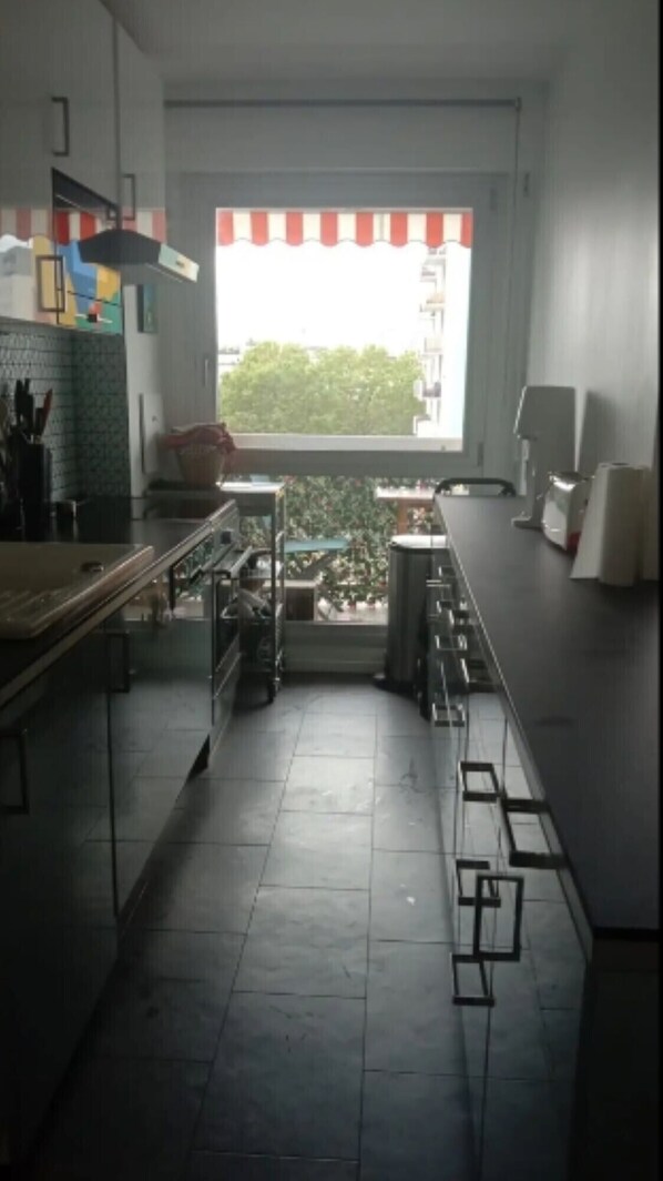 A narrow kitchen with a view of greenery outside, a window with a striped blind, and a tiled floor.