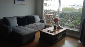 A living room with a grey sofa, a wooden coffee table, and a large window with a view of buildings and greenery.