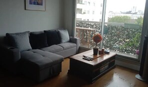 A living room with a grey sofa, a wooden coffee table, and a large window with a view of buildings and greenery.