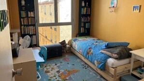 A small, well-lit bedroom with a single bed, a desk, a chair, and a bookshelf.