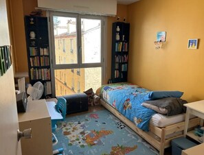 A small, well-lit bedroom with a single bed, a desk, a chair, and a bookshelf.