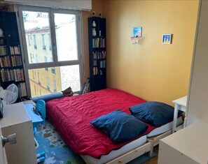 A small bedroom with a red bed, a bookshelf, a window with a view of buildings, and a white desk.
