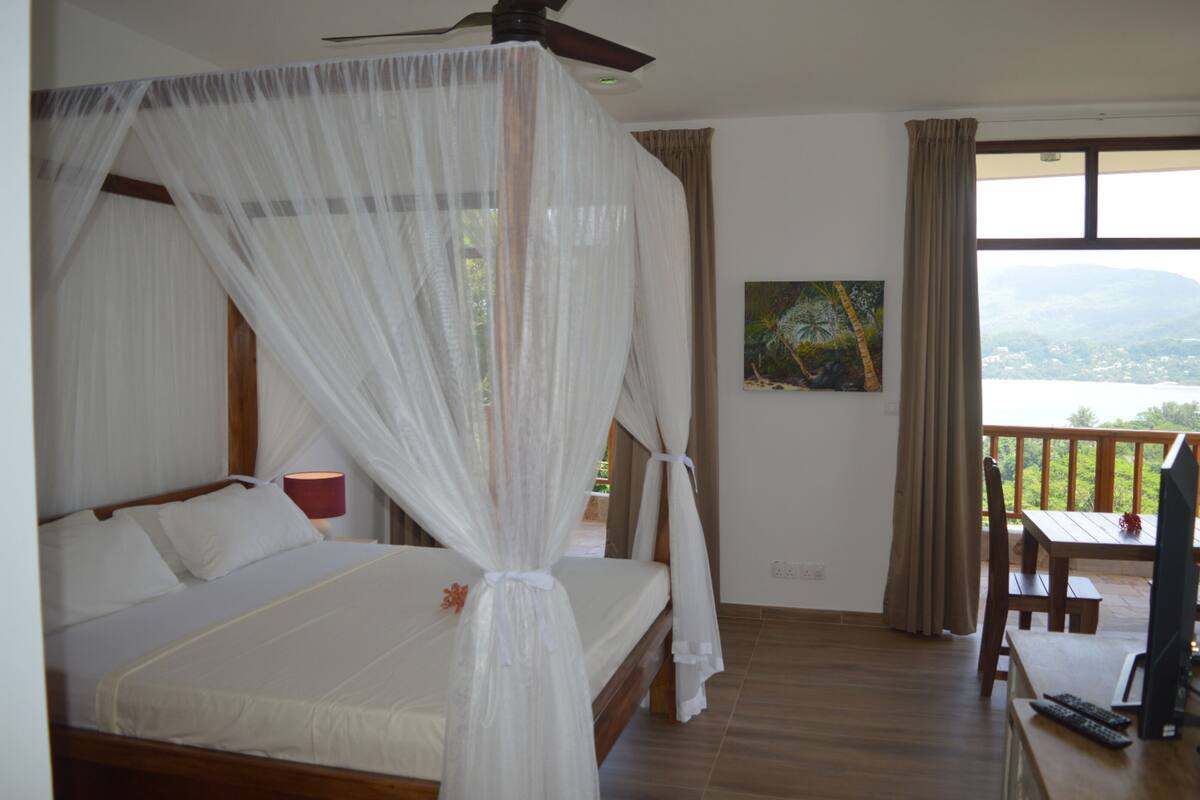 A bedroom with a bed, a ceiling fan, a painting, and a balcony with a view.