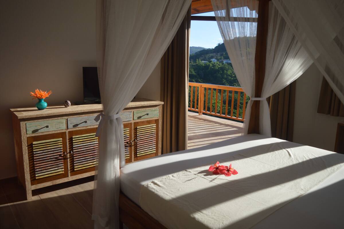 A bedroom with a bed, curtains, a wooden dresser, and a view of a balcony.