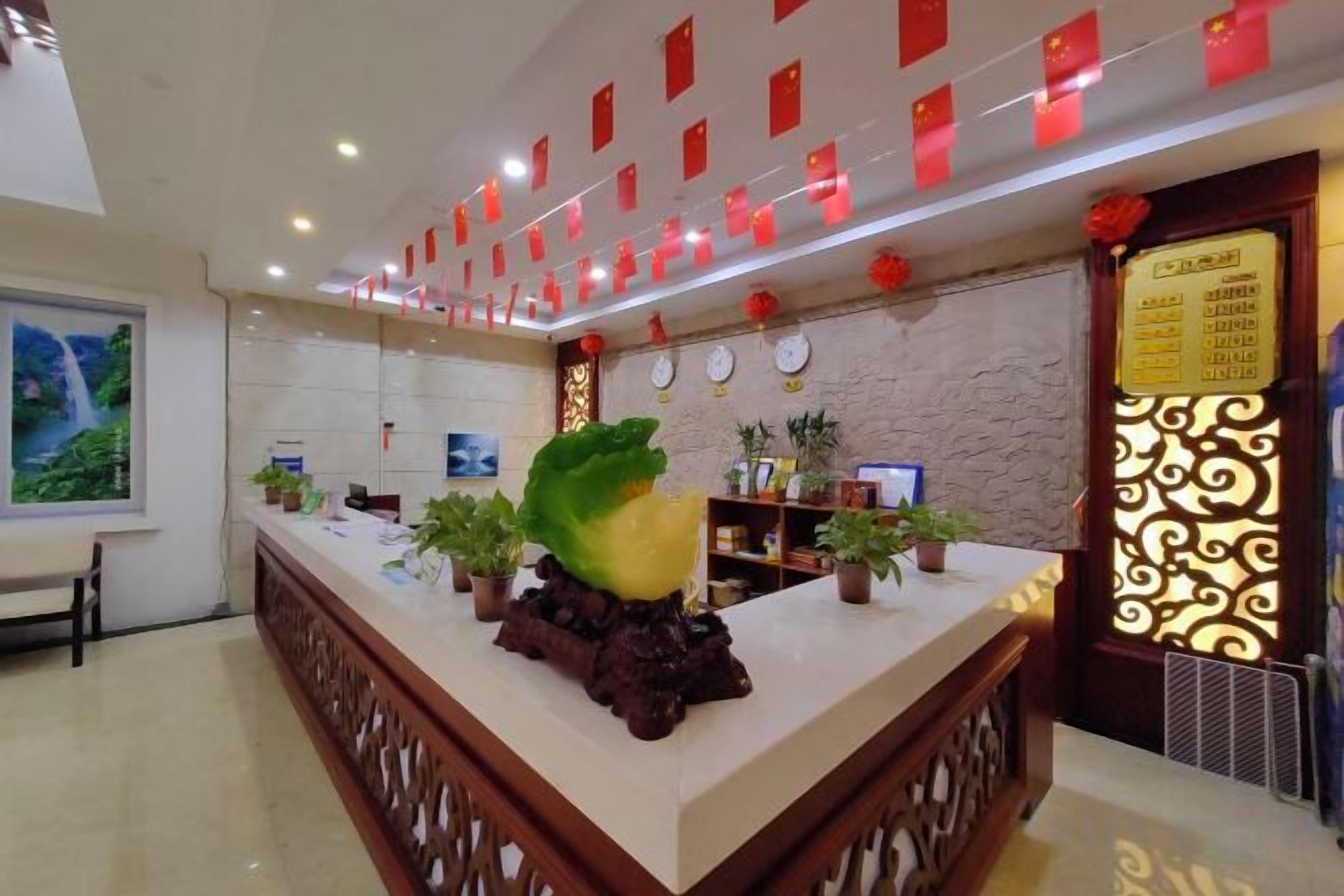 A hotel reception area with a reception desk, potted plants, a clock, and a wall with a decorative pattern.