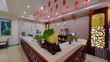A hotel reception area with a reception desk, potted plants, a clock, and a wall with a decorative pattern.