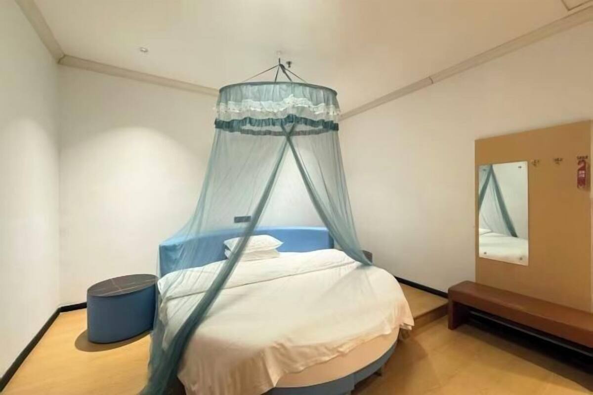 A bedroom with a bed under a mosquito net, a round blue ottoman, a bench, and a mirror on the wall.