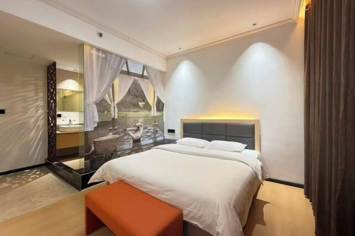 A modern hotel room with a bed, a bench, a mirror, and a bathroom visible through a glass partition.