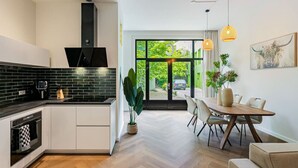 A modern kitchen with a black backsplash, white cabinets, and a built-in oven. There is a dining area with a wooden table and chairs, and a large window offering a view of greenery outside.