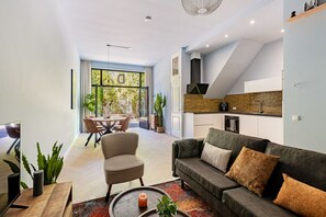 Uma sala de estar moderna com sofá, poltrona e mesa de centro, uma área de jantar com mesa e cadeiras, e uma cozinha com painel de parede de tijolos e armários brancos.