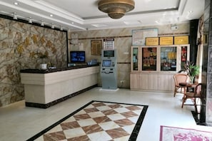 A reception area with a marble wall, a check-in counter, a self-service kiosk, a vending machine, and a seating area with a checkered floor.