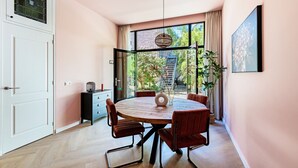 Uma área de refeições moderna com uma mesa redonda, cadeiras vermelhas e vista para um jardim através de grandes janelas.