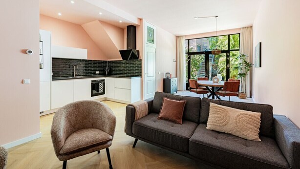 Uma sala moderna com sofá e poltrona, área de jantar com mesa e cadeiras, e cozinha com painel atrás do fogão preto.