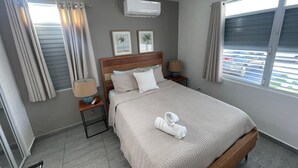 A hotel room with a bed, two bedside tables, a nightstand, an air conditioner, and a window with curtains. - Serenity House on Isla Verde Beach (Carolina)