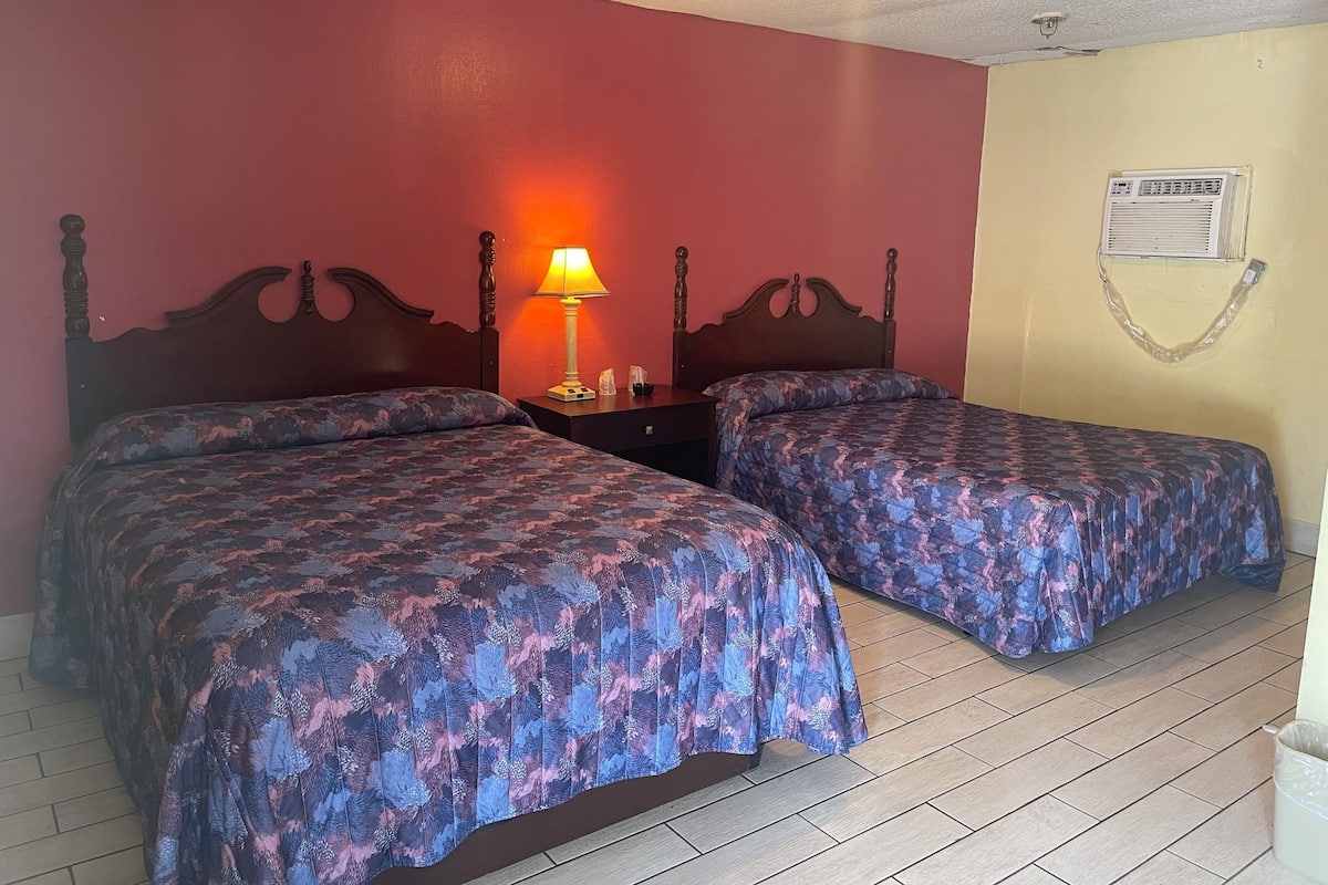 A room with two beds, a nightstand with a lamp, and a wall-mounted air conditioner.