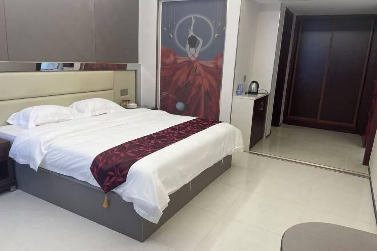 Habitación de hotel con cama, mesita de noche, televisor montado en la pared, puerta con espejo y una pequeña mesa con hervidor.