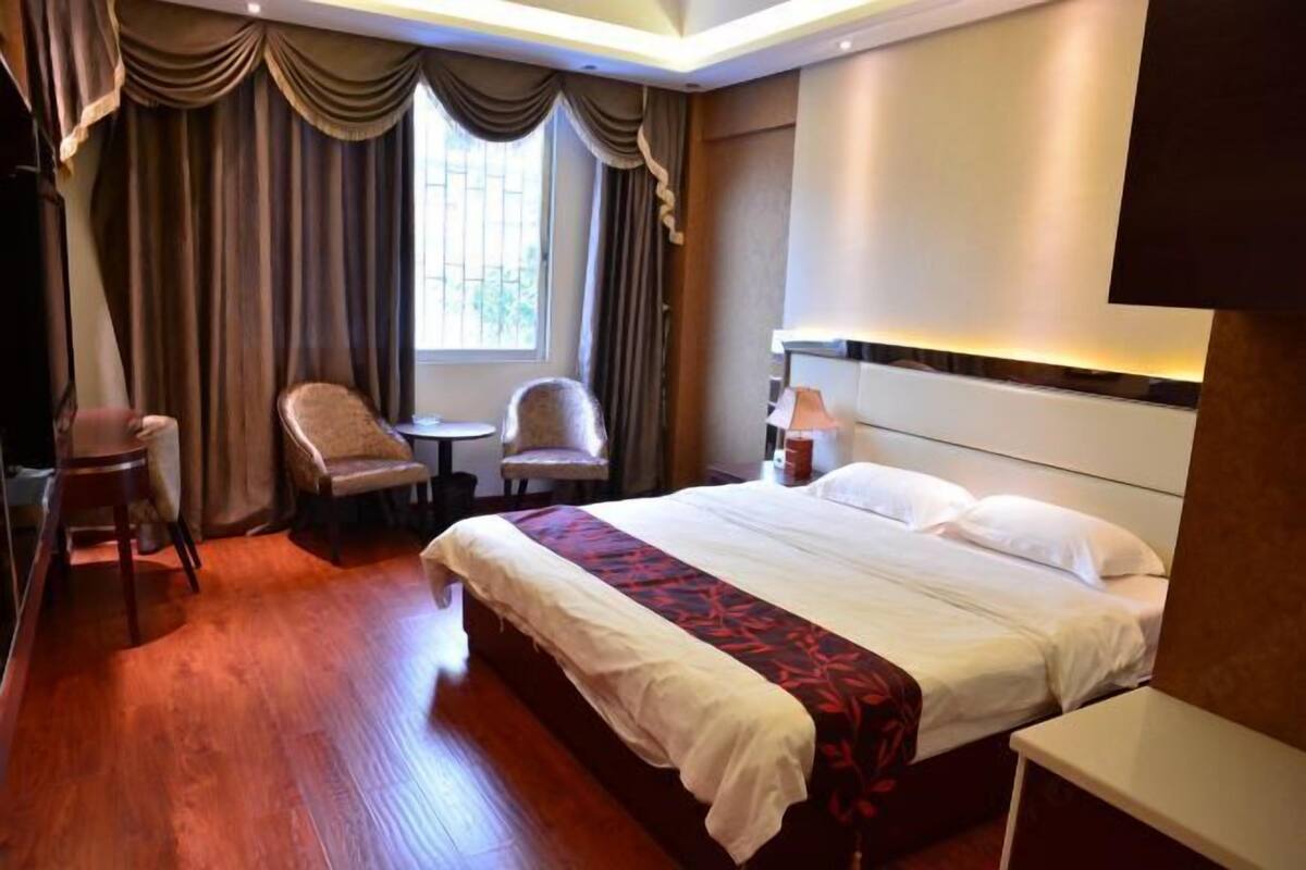 Una habitación de hotel con una cama grande, dos sillas, una mesita y un ventanal con cortinas.