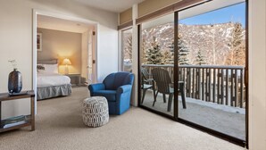 A hotel room with a bed, a blue armchair, a patterned ottoman, and a large sliding glass door leading to a balcony with a view of snow-covered trees and mountains. - Lichenhearth 25 (Snowmass Village)