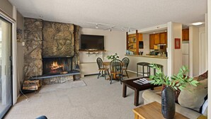 A living room with a stone fireplace, a dining area, and a kitchen in the background. - Lichenhearth 25 (Snowmass Village)