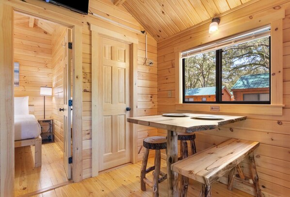 A wooden cabin interior with a bed, a table with stools, a window with a view of trees, and a door.