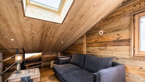 Un acogedor cuarto ático con techo de madera, un sofá gris oscuro, una mesita con lámpara y un televisor.