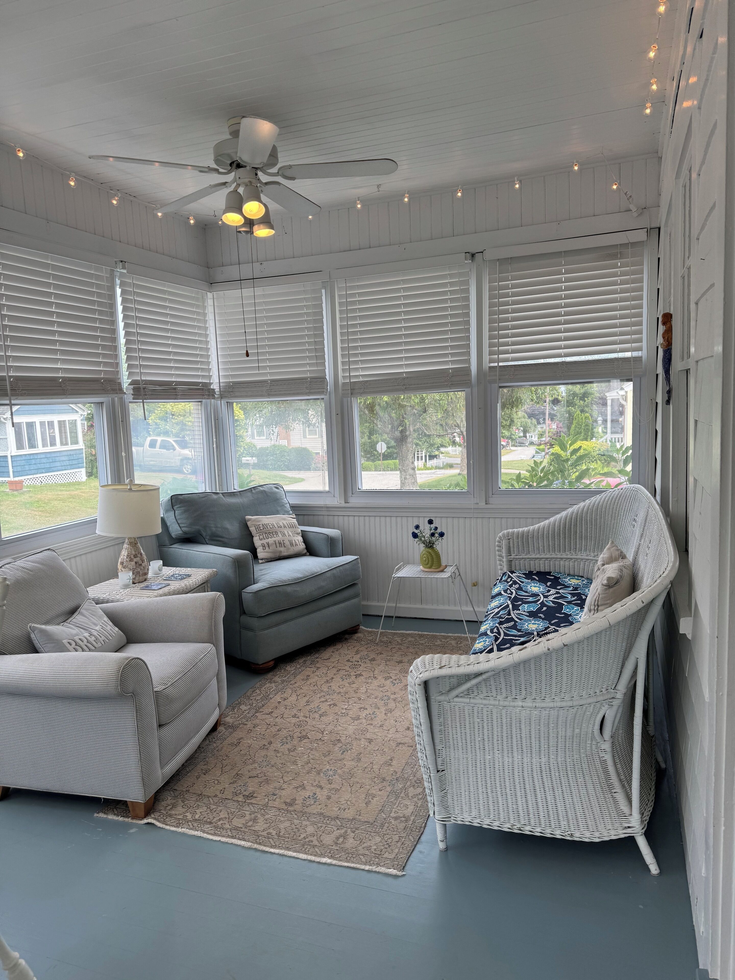 A room with a ceiling fan, two grey sofas, a wicker chair, a small table with a lamp, and a view of the outside.