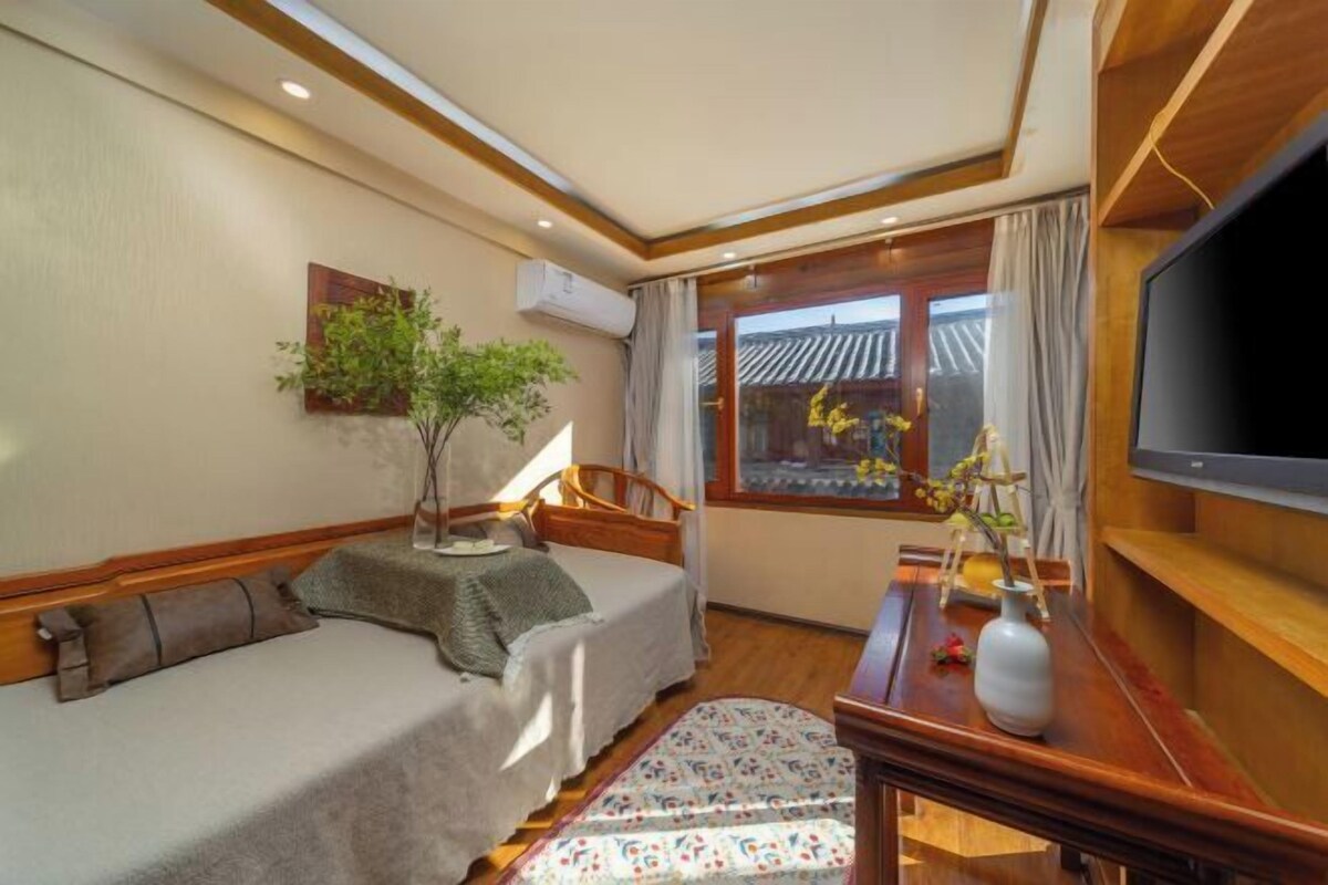 A hotel room with a bed, a desk with a TV, a window with curtains, a plant, and a wall-mounted air conditioner.