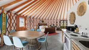 Un espacio habitable acogedor y de concepto abierto, con techo de madera, un comedor con mesa y sillas redondas, una cocina con estufa, y una cama con cabecera.
