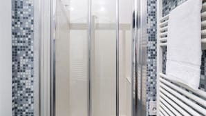 Ducha con mampara de vidrio, pared con mosaico de azulejos y una toalla blanca colgada en un perchero.