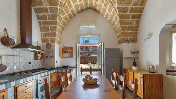 Una cocina con una larga mesa de madera, salpicadero de azulejos y techo abovedado de piedra.