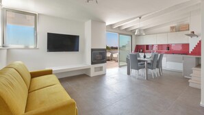 A modern living room with a yellow sofa, a flat-screen TV mounted on the wall, and a dining area with a table and chairs.