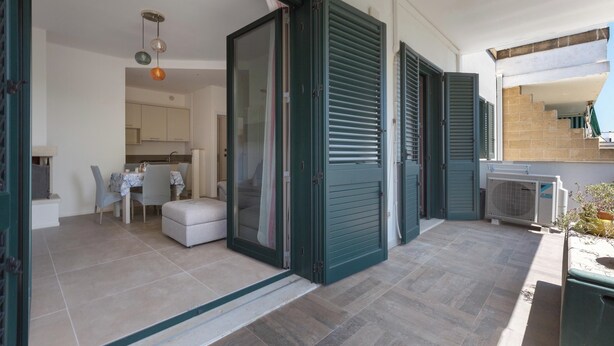 Una puerta corredera abierta da a un comedor con una mesa y sillas, una cocina visible a través de la puerta de vidrio, y un pequeño espacio al aire libre con una mesa y sillas.