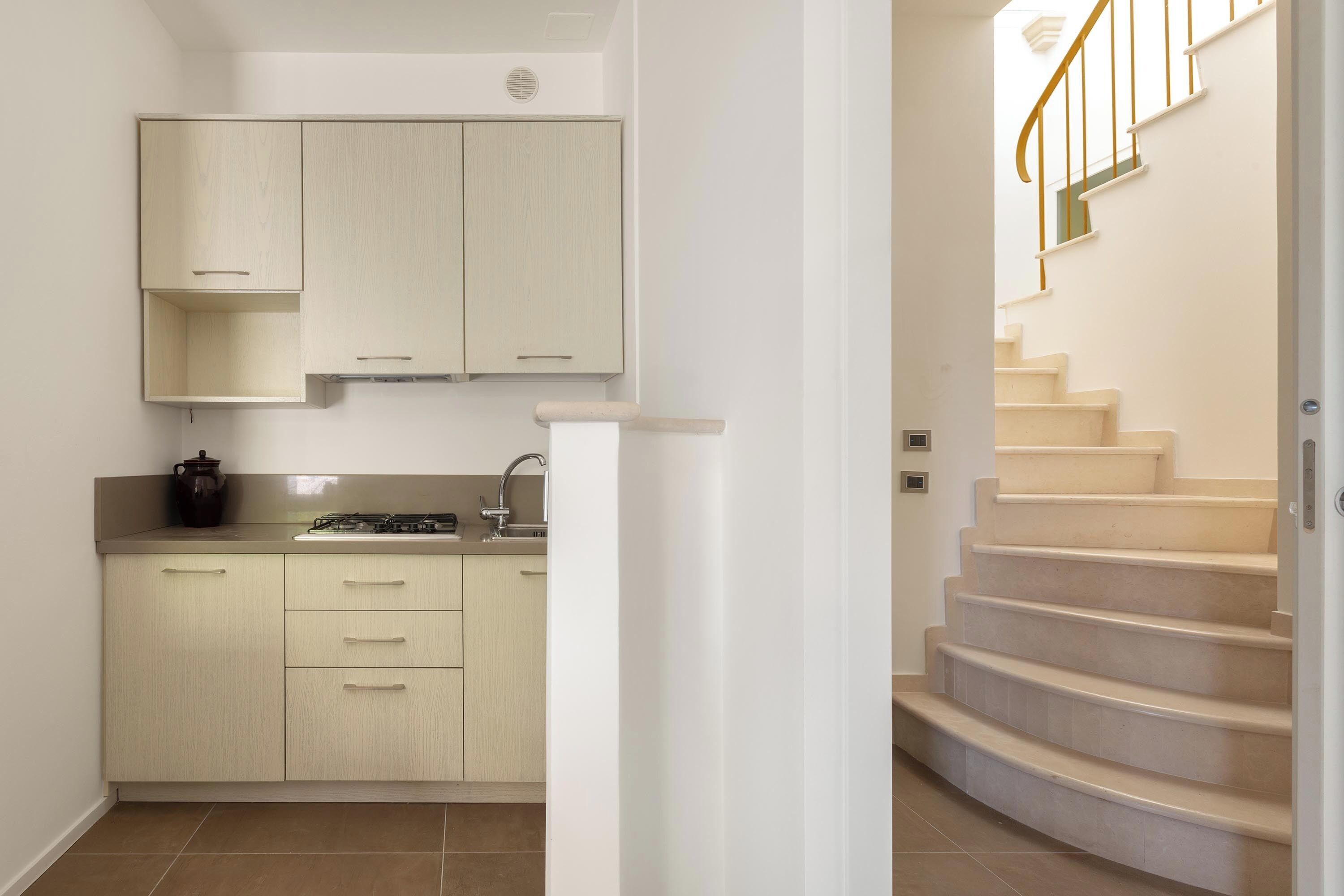 A kitchen with light-colored cabinets, a sink, and a stove. There is a staircase with a handrail leading to an upper floor.