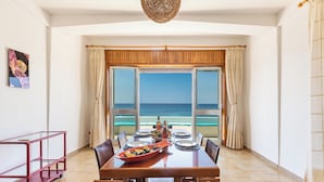 Comedor con una mesa dispuesta para cuatro, un carrito metálico rojo y vistas al océano a través de amplios ventanales.