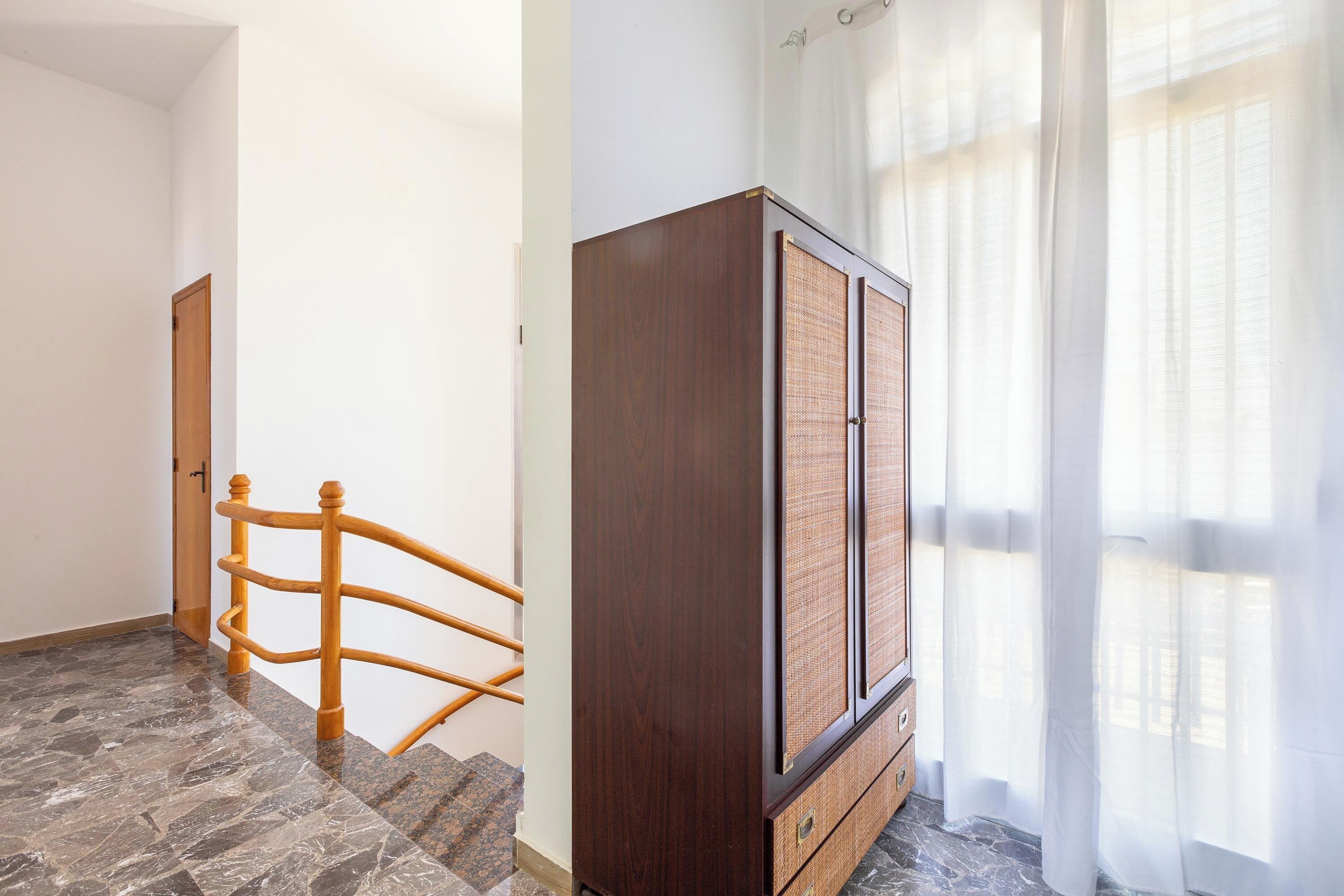 Una escalera con barandilla de madera que lleva a una habitación con un armario de madera oscura y una ventana con cortinas livianas.