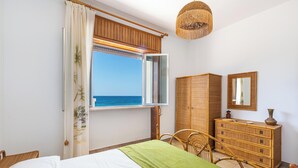 Un dormitorio con una cama, un tocador, una silla, un ventanal con vista al mar y una lámpara colgante.