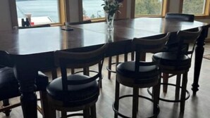A large dining table with six chairs, a vase with flowers, and a view of a lake and mountains through large windows.