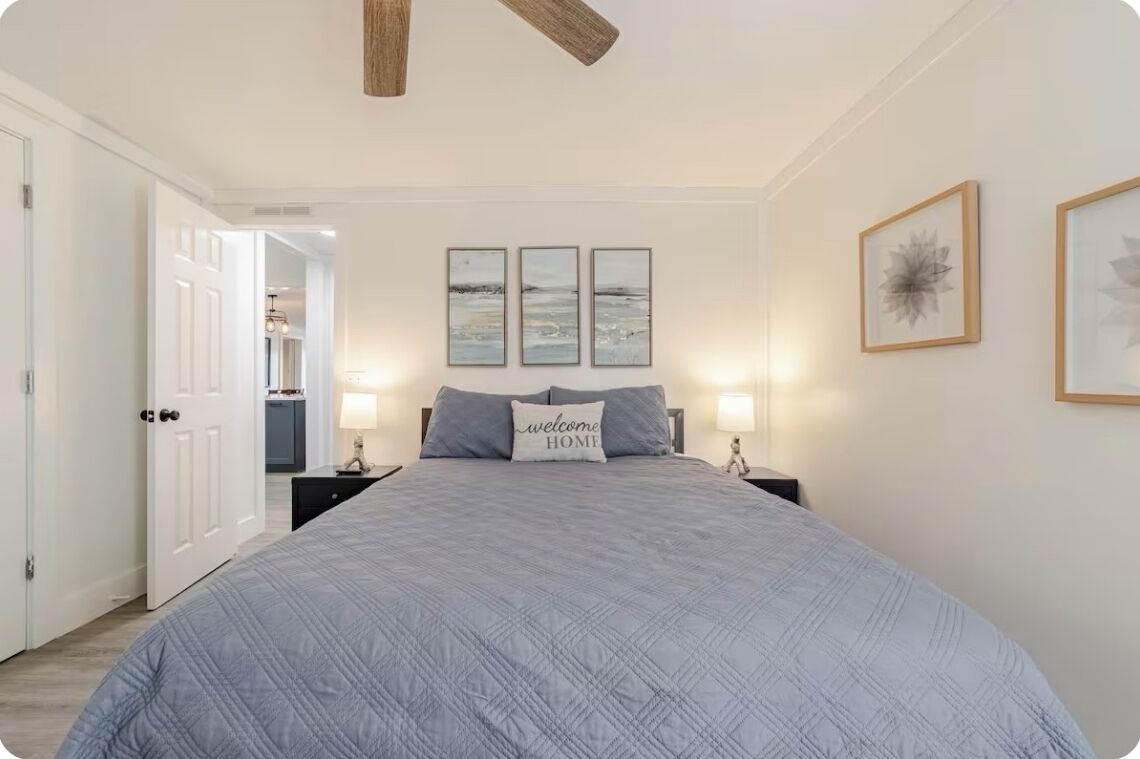 A bedroom with a large bed, a ceiling fan, three framed artworks on the wall, and a 'welcome home' pillow.