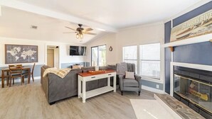 Un salón amplio con un sofá gris, una mesa de centro blanca y un sillón gris. Hay una chimenea con un televisor montado encima, y un mapa del mundo en la pared.