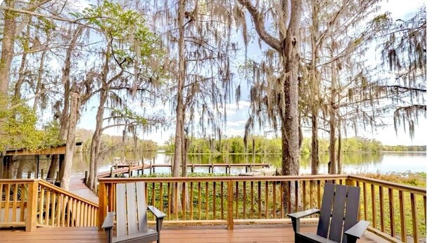 Una terraza de madera con dos sillas Adirondack frente a un lago, rodeado de árboles cubiertos de musgo español.