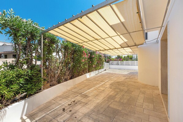 A covered patio area with tiled flooring, a white wall, and a row of plants.