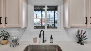 Una cocina con armarios blancos, un lavamanos de acero inoxidable, una ventana con vistas a edificios, una planta pequeña y un dispensador de jabón.