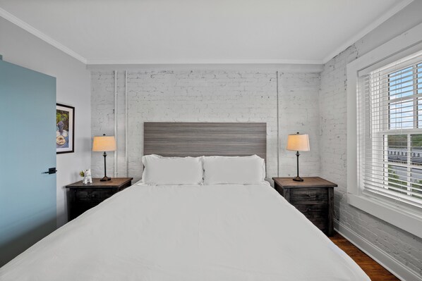 Une chambre à coucher bien aménagée, avec un grand lit, deux tables de chevet avec des lampes, un mur blanc décoré d’un motif imitant des briques et une fenêtre avec des stores.