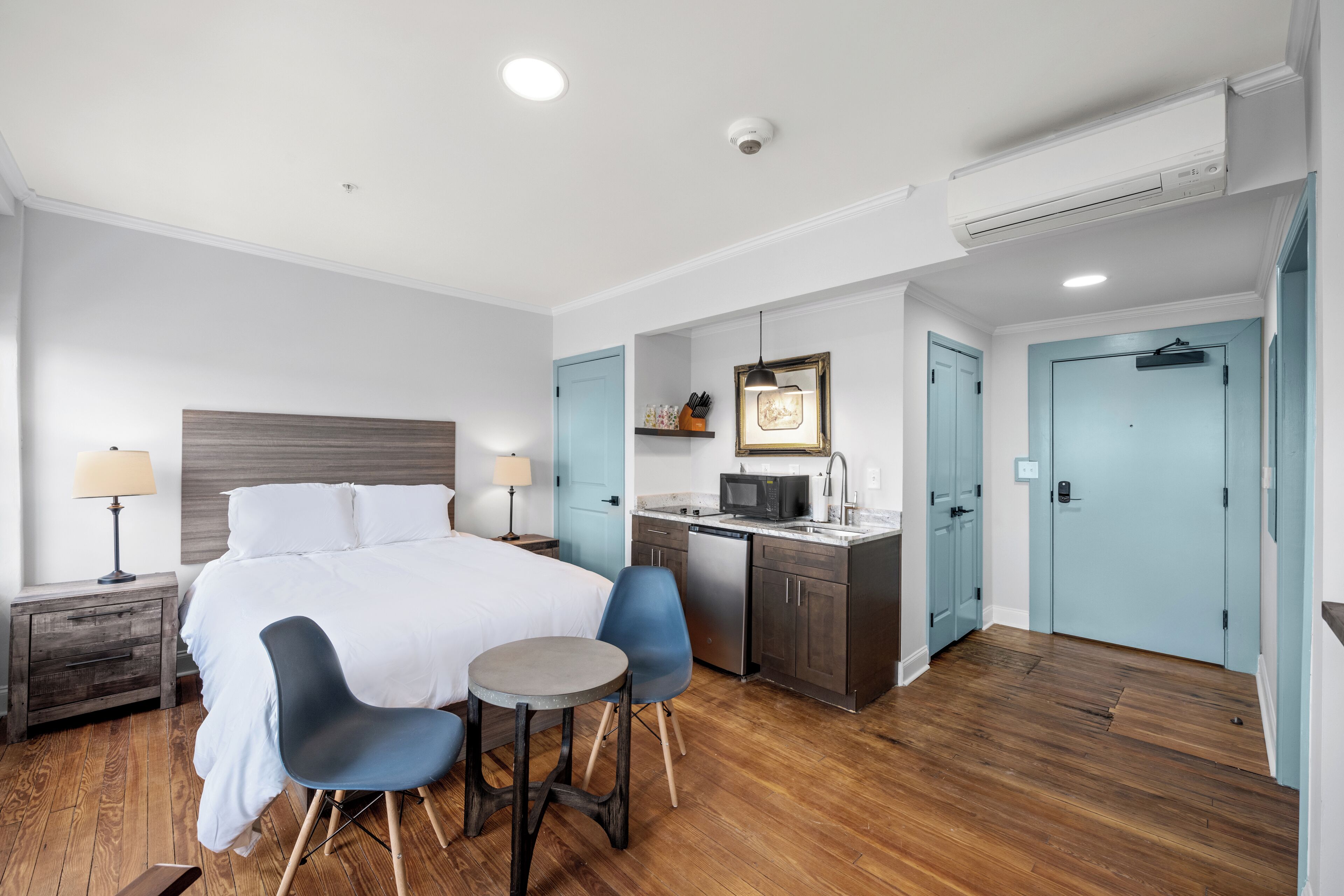 A hotel room with a bed, two chairs, a small table, a kitchen area, and a bathroom.