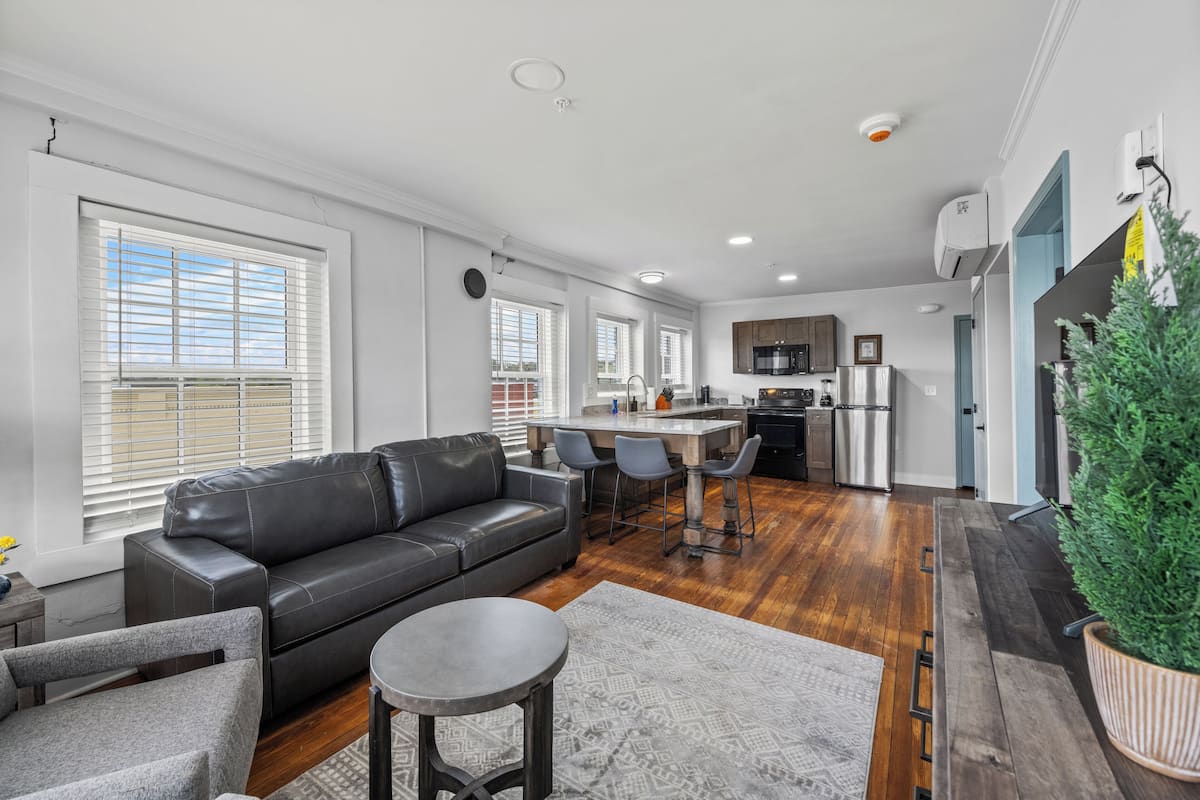 A modern living room with a black leather sofa, grey armchair, and a round coffee table. A kitchen area with a bar counter and stools is visible in the background.