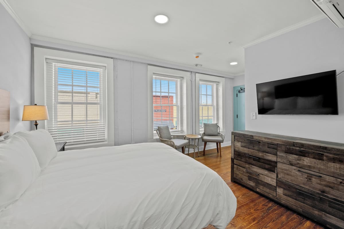 A hotel room with a large bed, a TV mounted on the wall, a wooden dresser, and a seating area with a table and chairs.