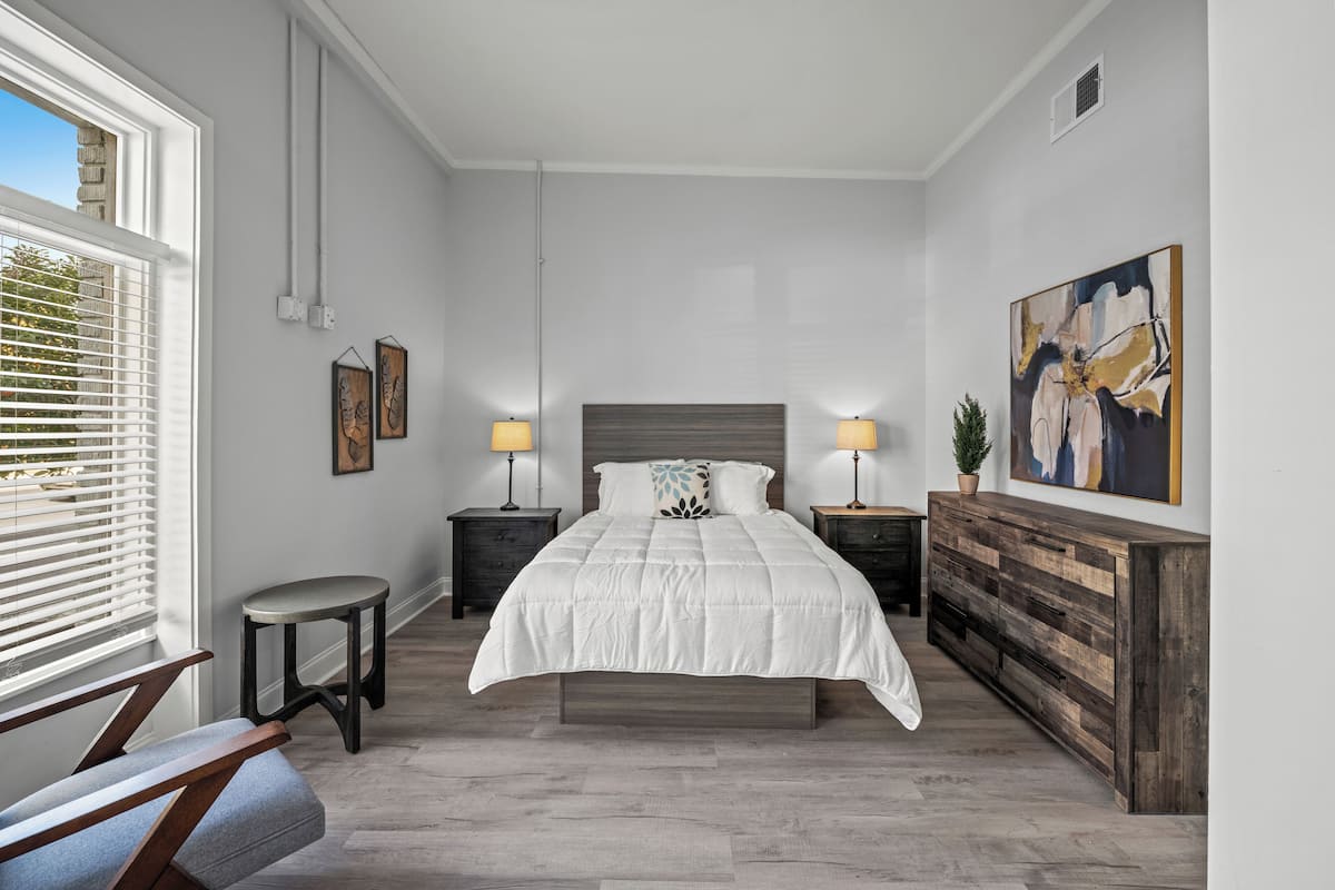 A bedroom with a large bed, two bedside tables with lamps, a small round table, a bench, and a wooden dresser.